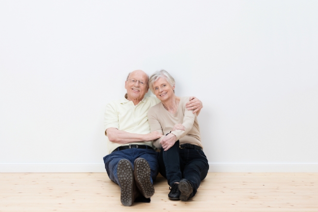 Loving elderly couple in their new home sitting side by side on the bare wooden floor smiling in satisfaction at having achieved their goal of a dream house